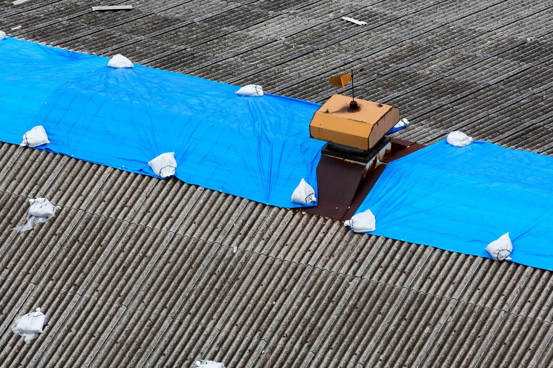 Storm damaged roof on house with a protective blue plastic tarp spread over hole in the shingles and rooftop.